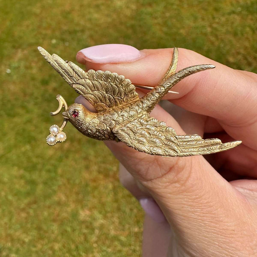 Victorian gold swallow brooch with pearl flower, symbolizing love and renewal, held in a hand against a grassy background.