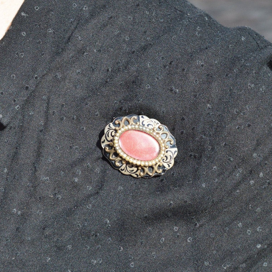 Victorian-style brooch with pink stone and intricate metalwork on a black fabric backdrop.