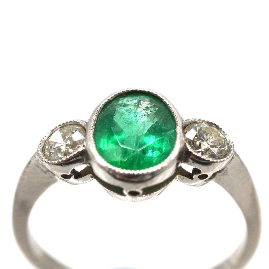 1950s platinum three stone bezel ring featuring a 1 carat emerald and bright diamonds, perfect for vintage engagement or anniversary.