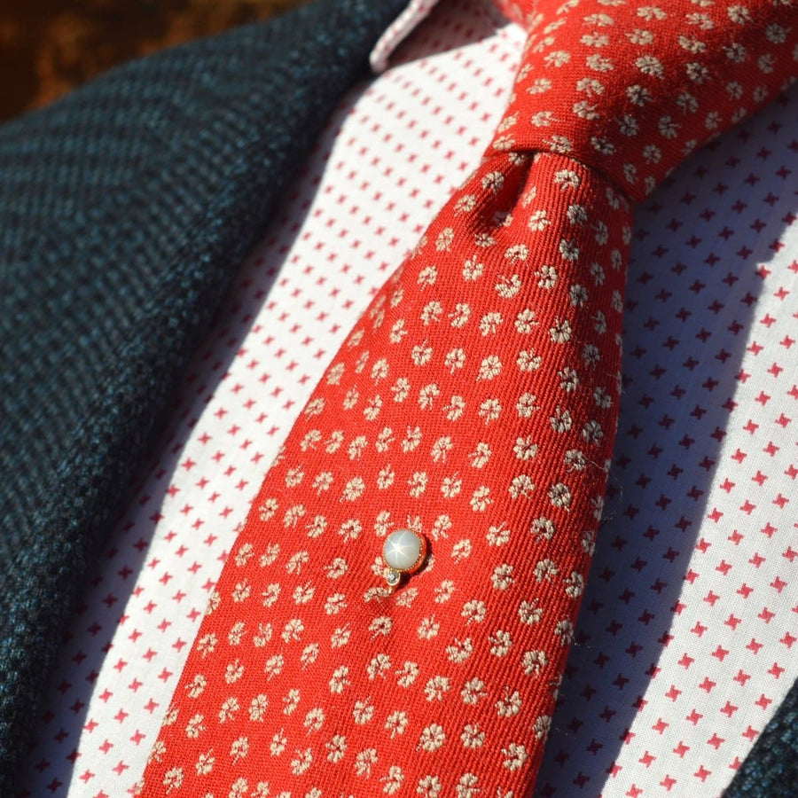 Close-up of a stylish red tie with floral patterns, featuring a pearl tie pin against a patterned shirt.