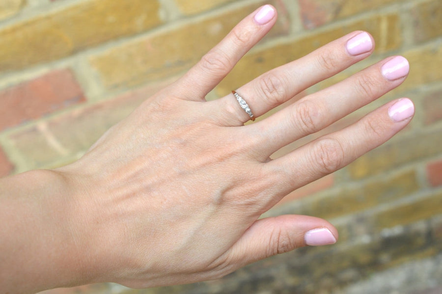 Elegant Art Deco 18ct gold and platinum three stone diamond ring showcased on a hand with pink nails against a brick background.
