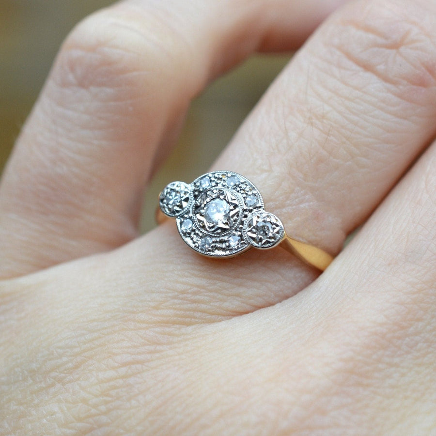 Art Deco 18ct gold and platinum three stone diamond halo ring, featuring intricate detailing and a central diamond.