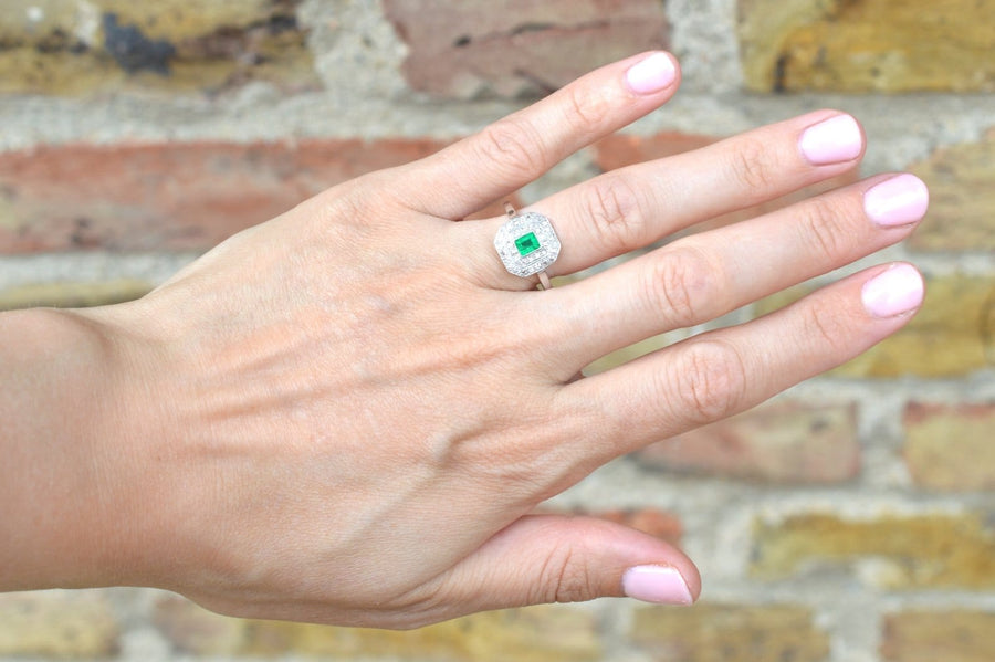 Art Deco 18ct white gold and platinum emerald and diamond ring worn on a hand against a brick background.