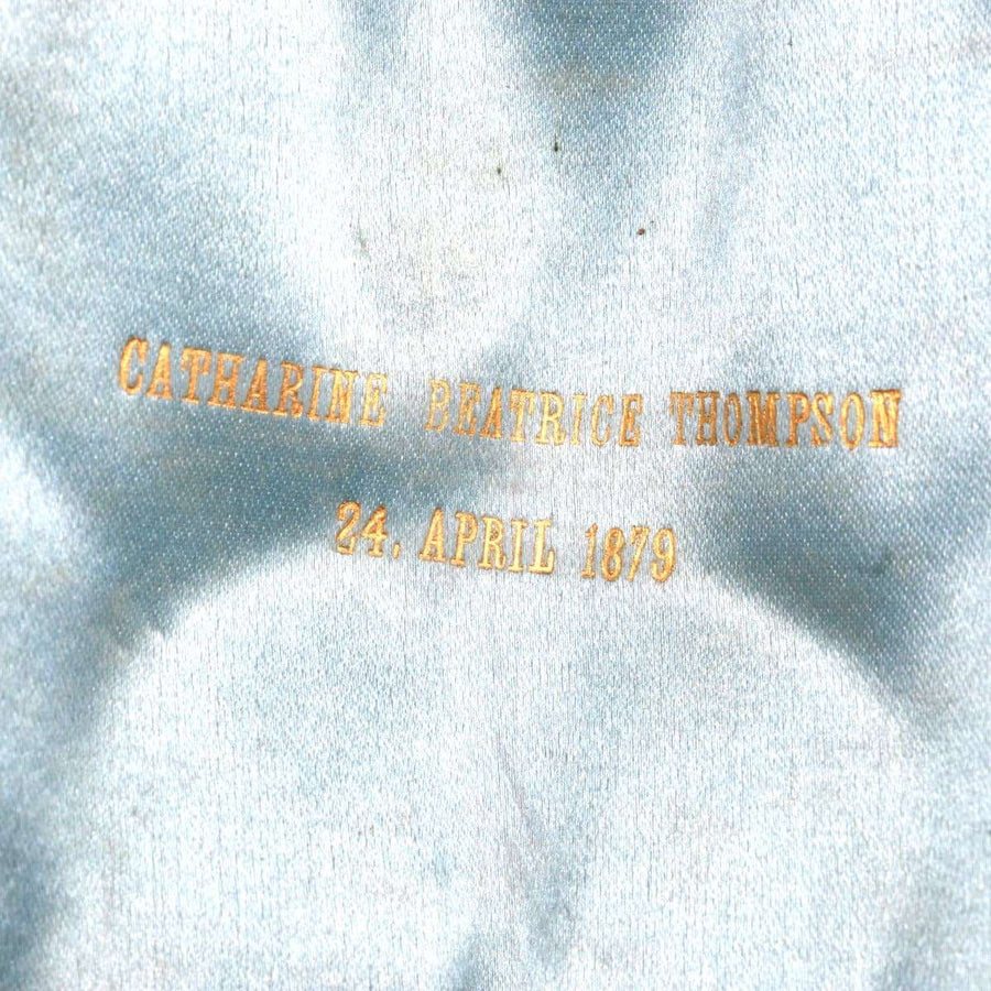 Gold inscription 'Catherine Beatrice Thompson 24th April 1879' on vintage satin background.