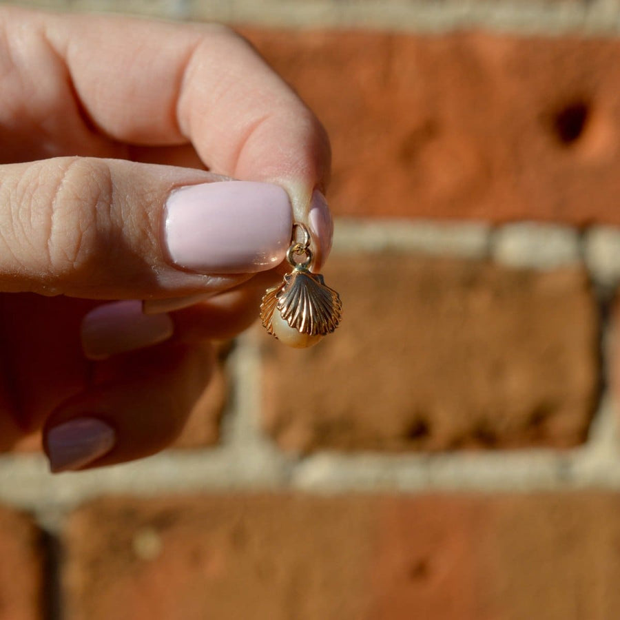 Vintage 9ct gold scallop shell charm pendant with imitation pearl held in hand against rustic brick background.